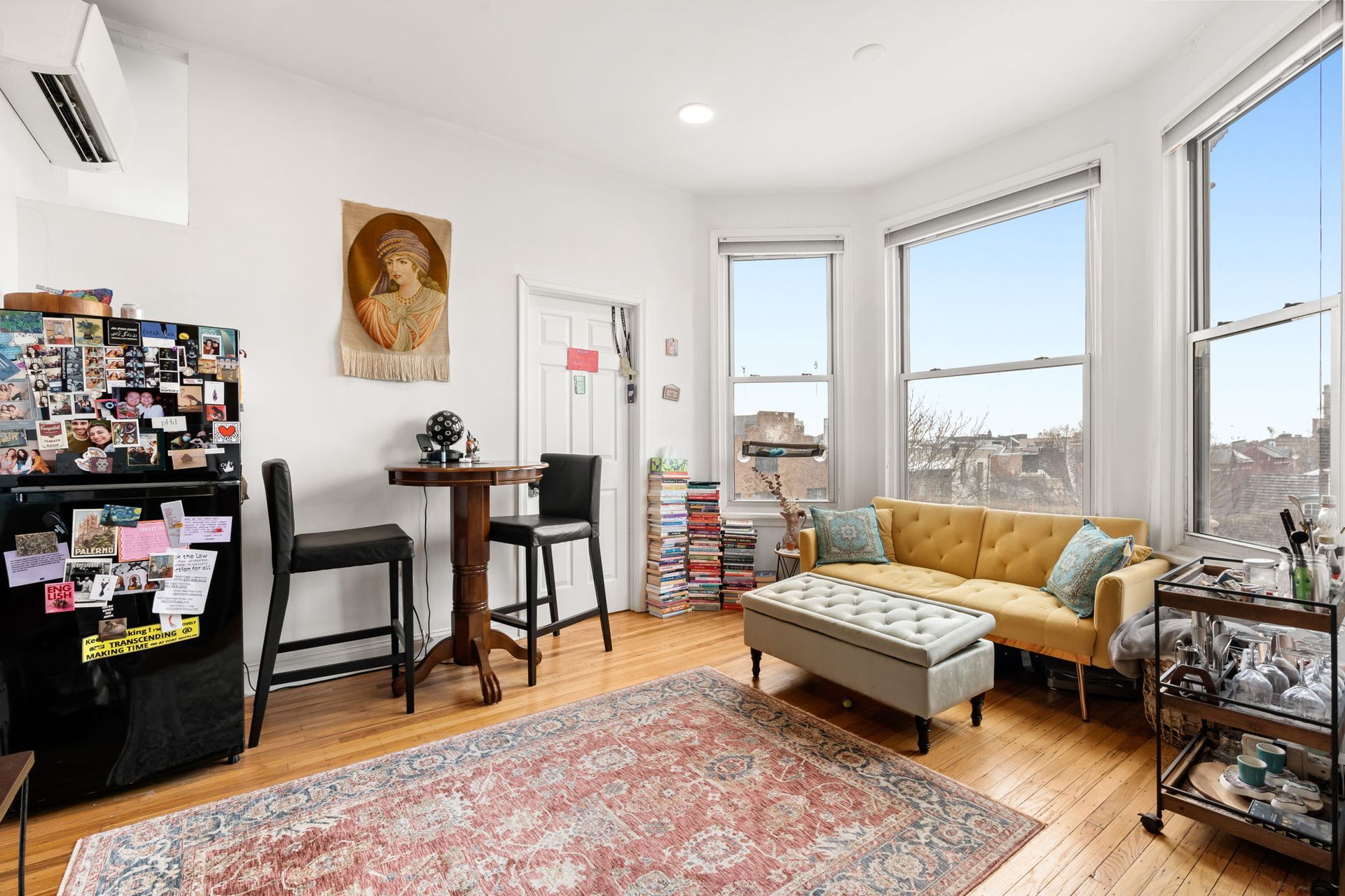 A cozy living area featuring a yellow sofa, an ottoman, a small table with two chairs, and a refrigerator covered in notes.