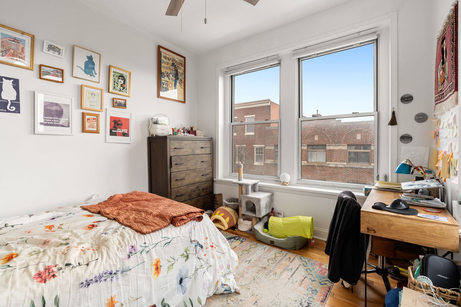 A bedroom with a bed featuring a floral quilt, a dresser, a desk by two windows, and a gallery wall of framed art.
