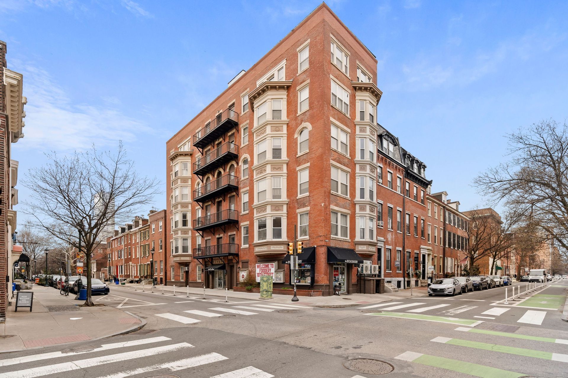 A multistory red brick apartment building with bay windows on a sunny corner in a city, next to a street intersection.
