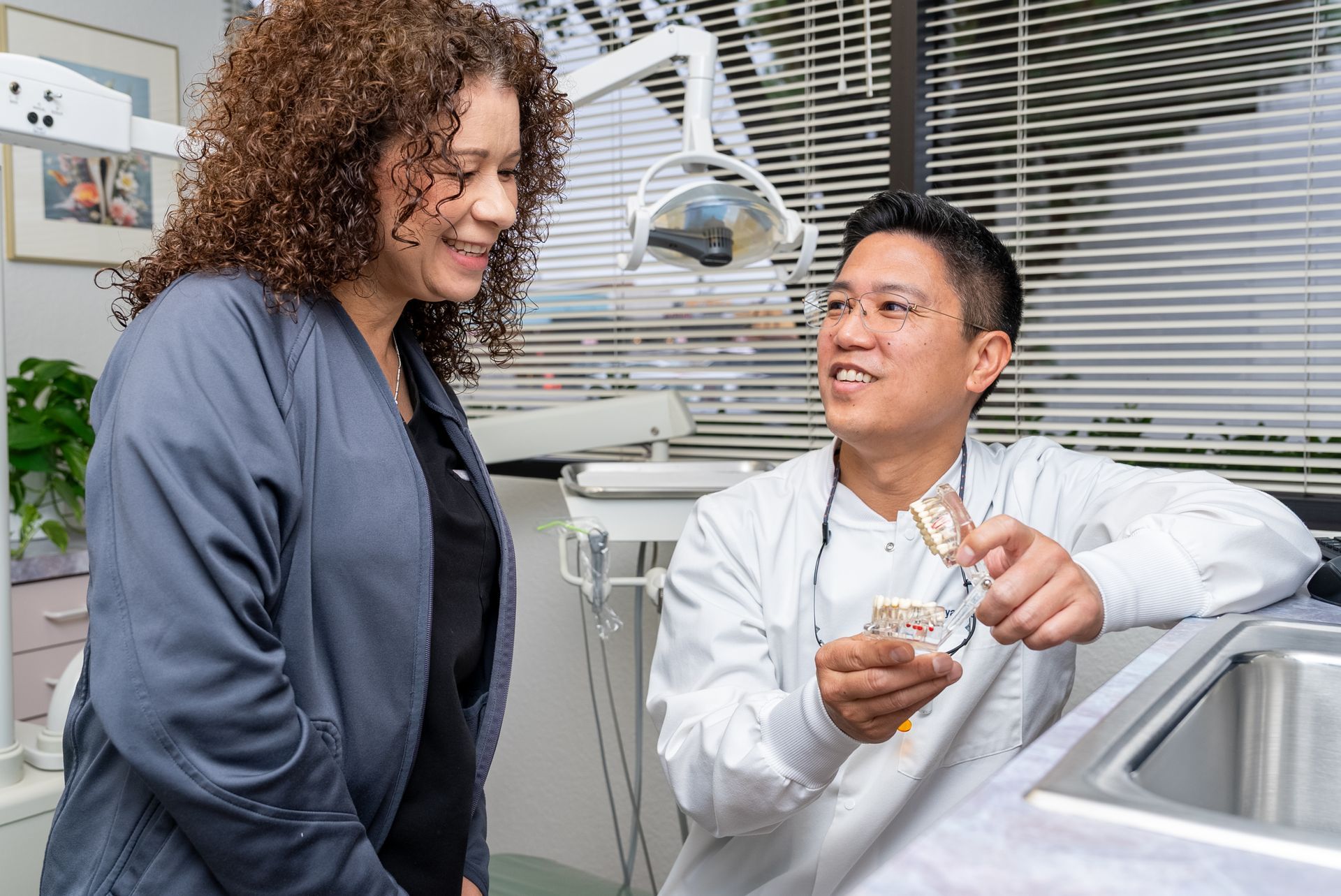 Dentist showing dental model to patient in office. White coat, gray scrubs, smiles.