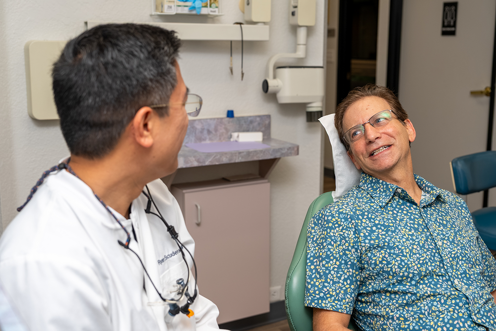 Dentist talking to patient in dental chair, examining patient's teeth. Bright setting.