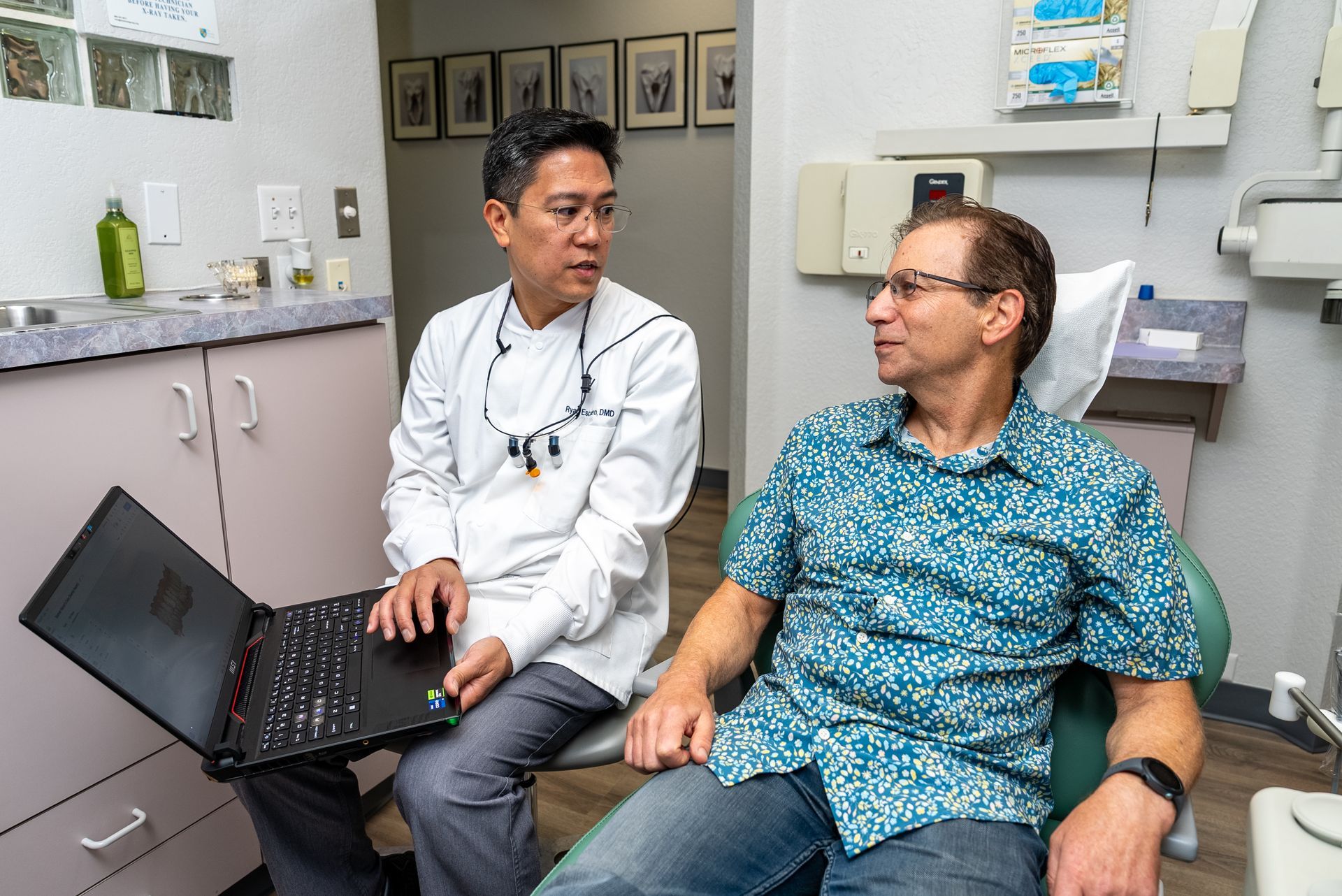 Dentist with laptop consults patient in dental chair, light-skinned interior.