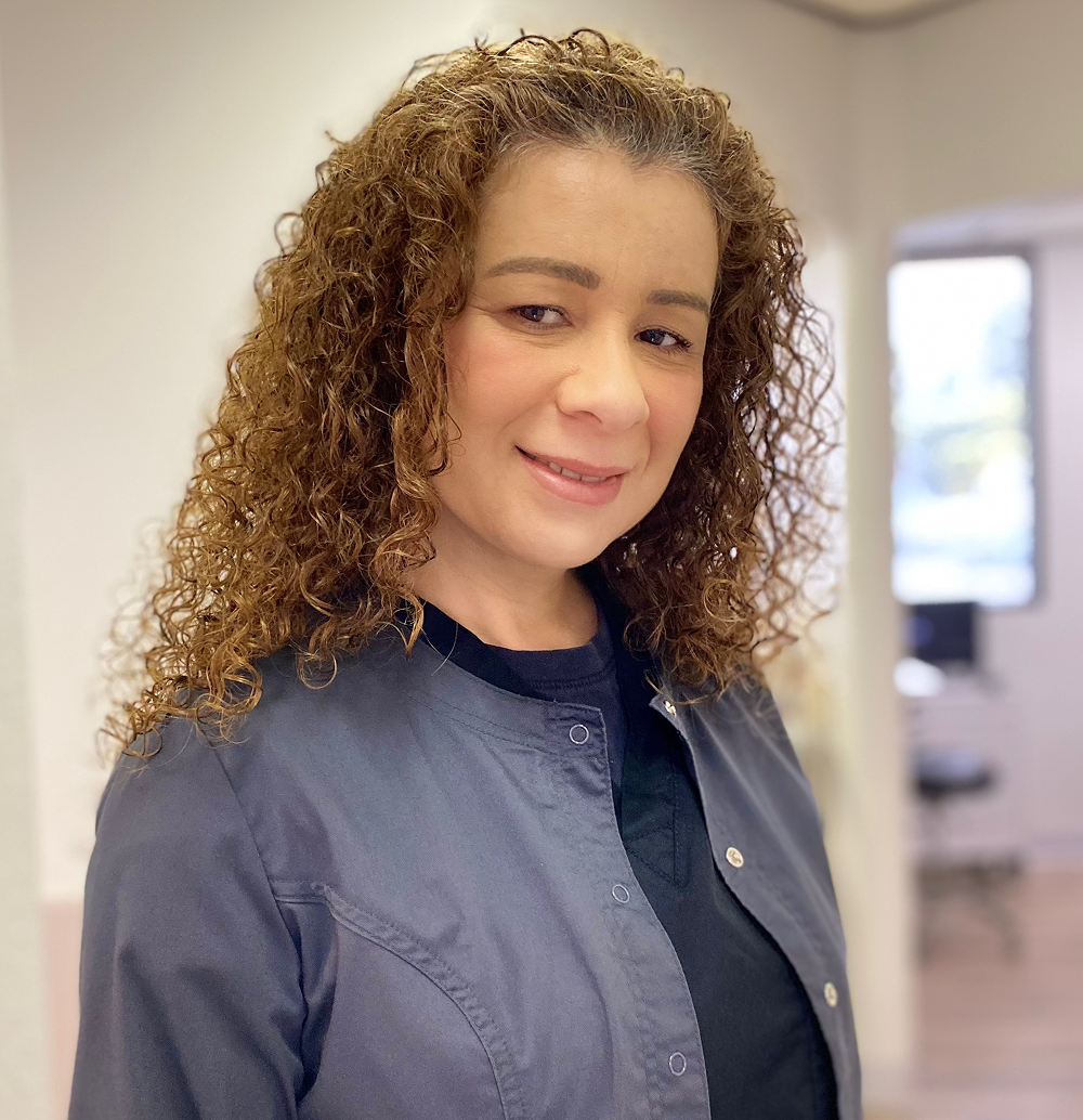 Woman with curly brown hair, wearing a gray jacket and black top, smiling in an office setting.