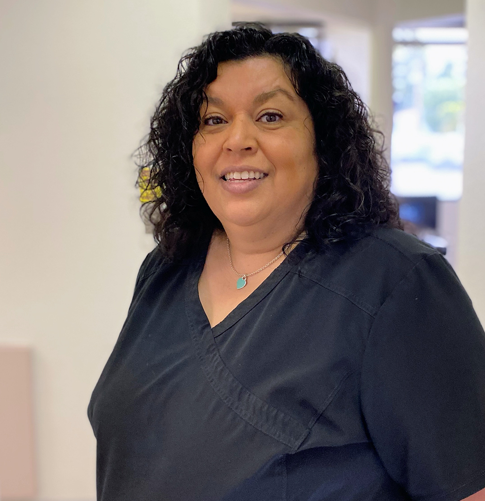 Woman with curly dark hair smiles, wearing a black v-neck shirt, indoors.
