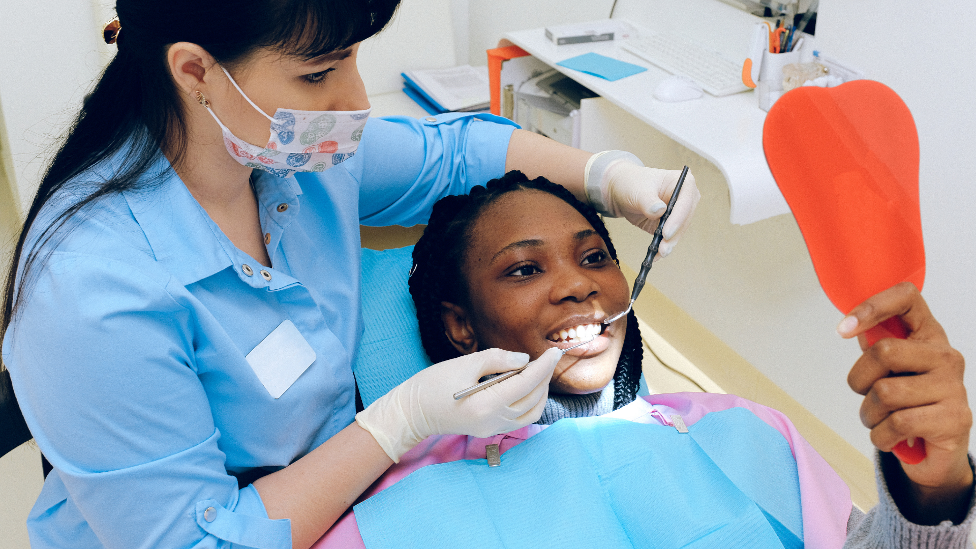 Dentist examining patient's teeth in a dental chair. The patient looks in a mirror.