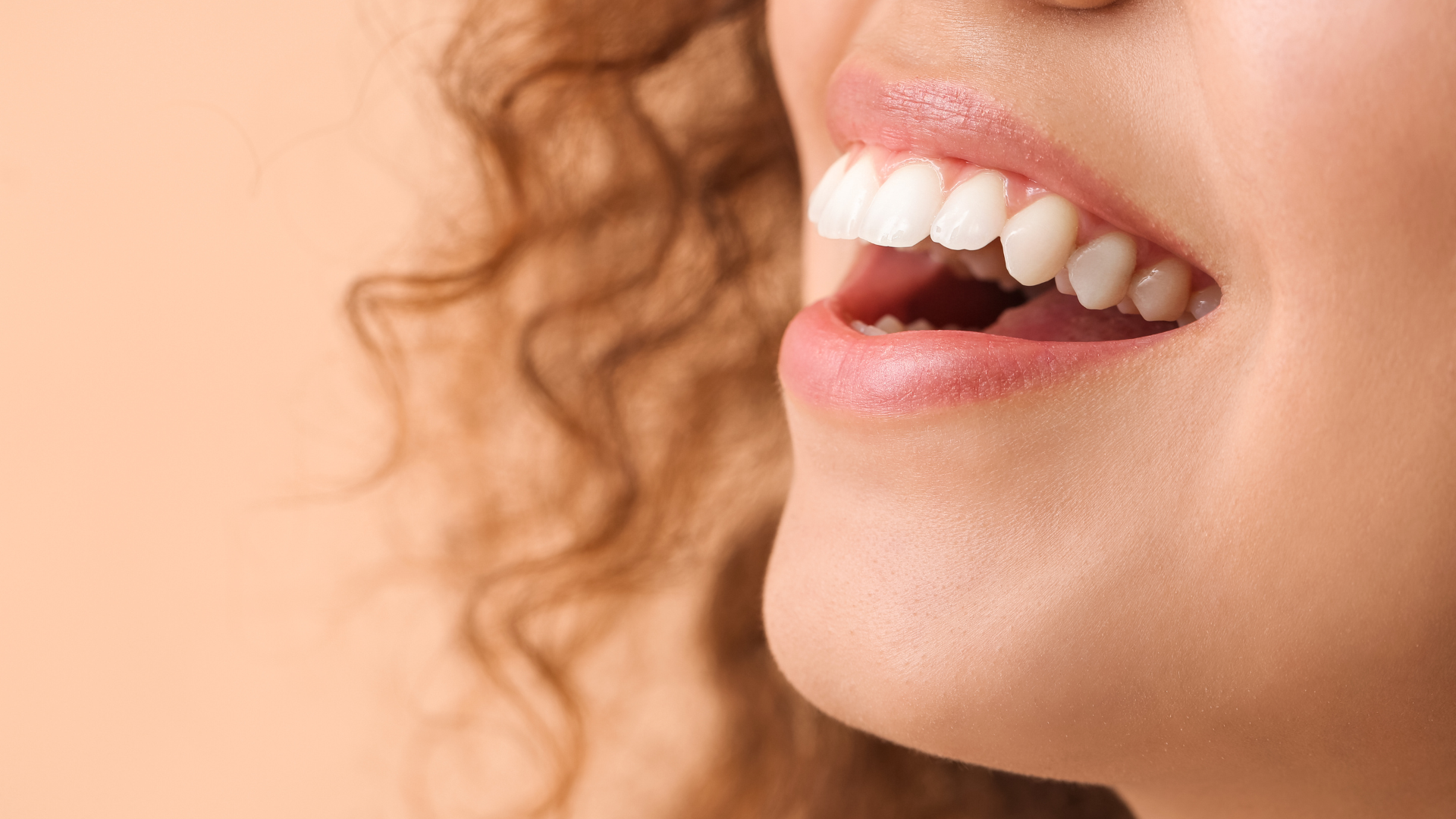 Close-up of a person's smiling mouth, showing white teeth against a peach background.