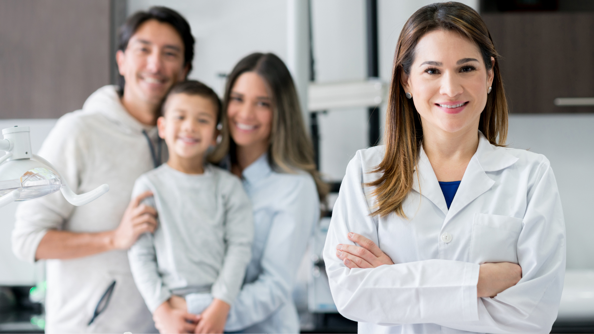 Dentist smiling, arms crossed, with a family in the background. Family includes two adults and a child.