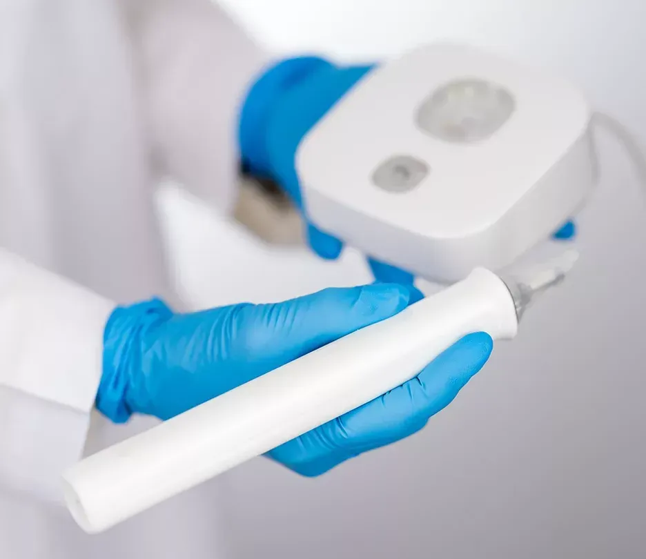 Hands in blue gloves holding a white medical device and accompanying base against a white backdrop.