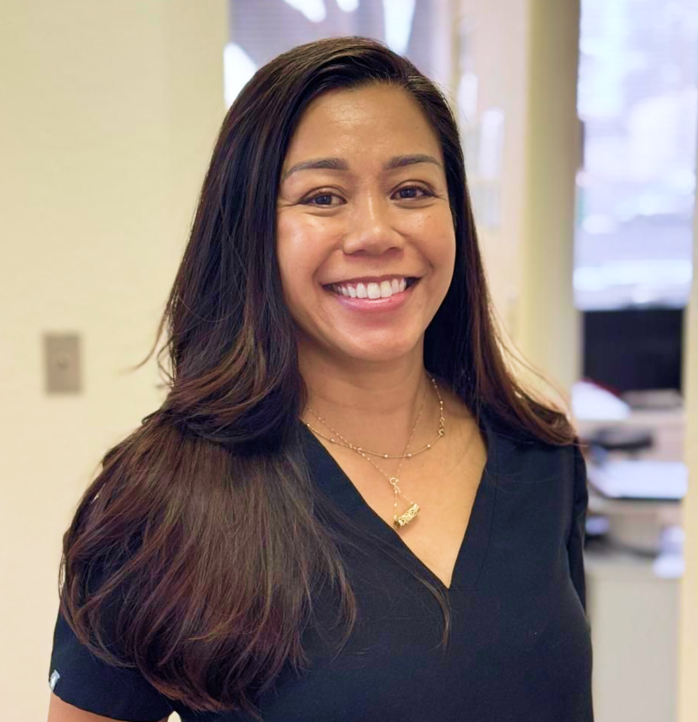 Woman in black top smiles, looking at the camera. She has long, dark hair, and a gold necklace.