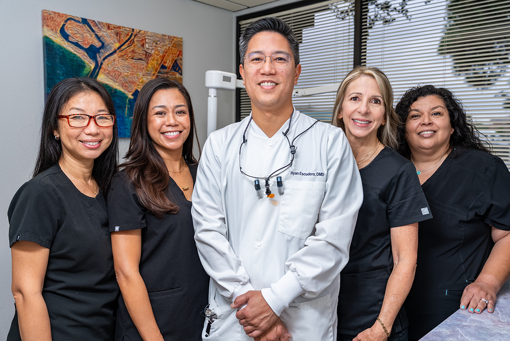 Dental office staff smiling, posing for a group photo; a colorful painting and window in the background.
