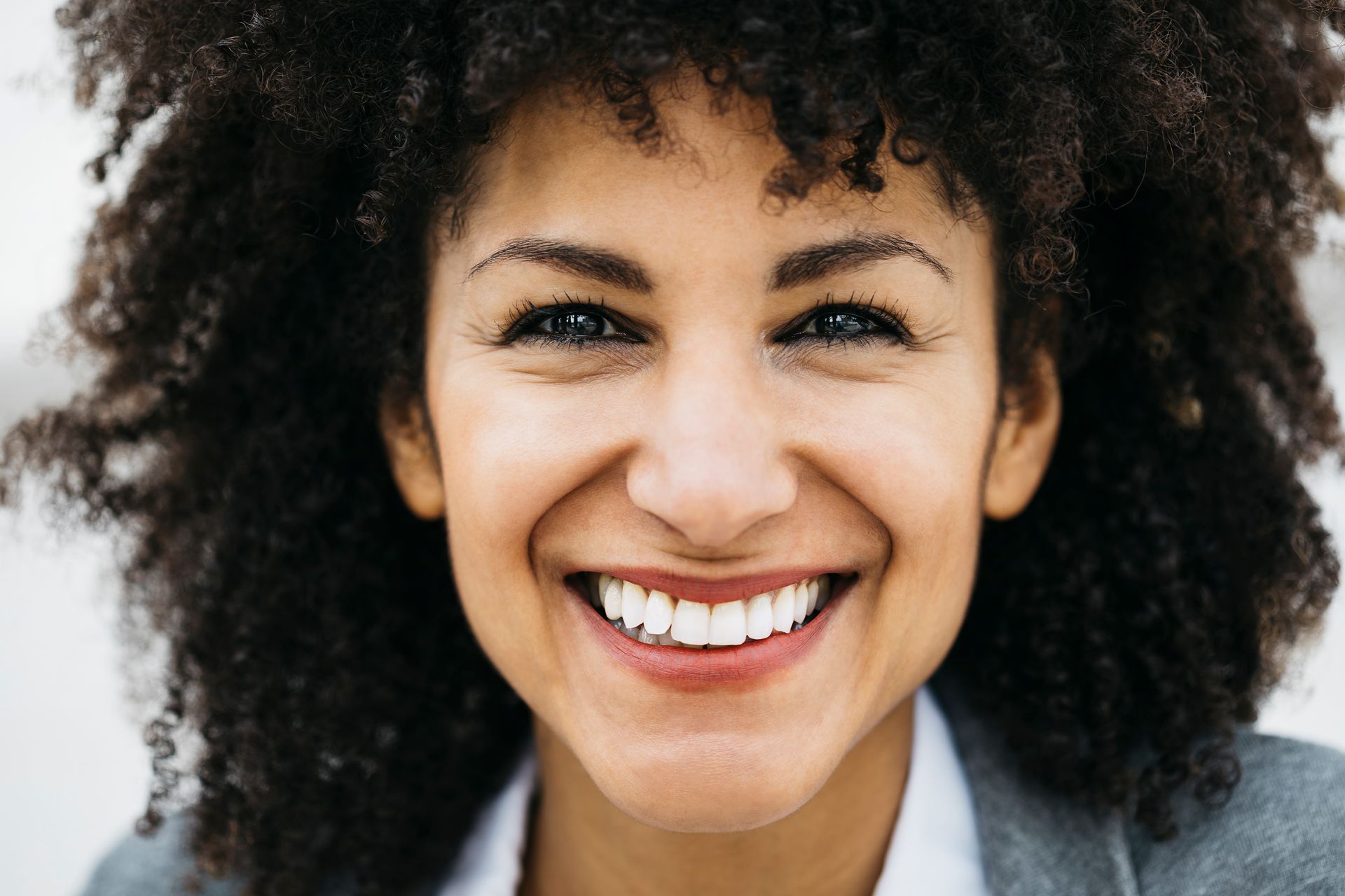Woman with curly hair smiles widely, showing teeth; blurred background.