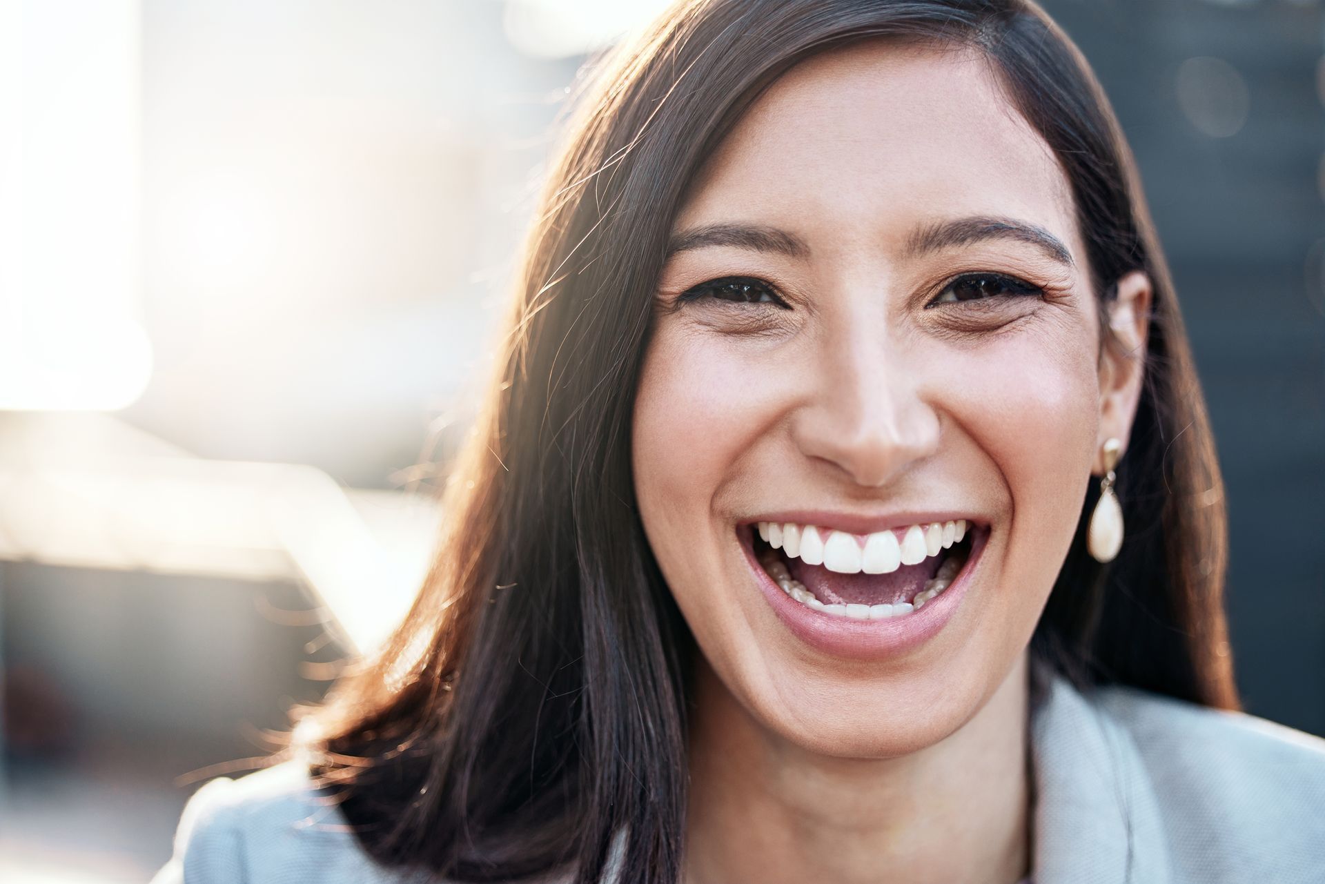 Woman laughing, wearing a light-colored blazer, with sunlight behind her.