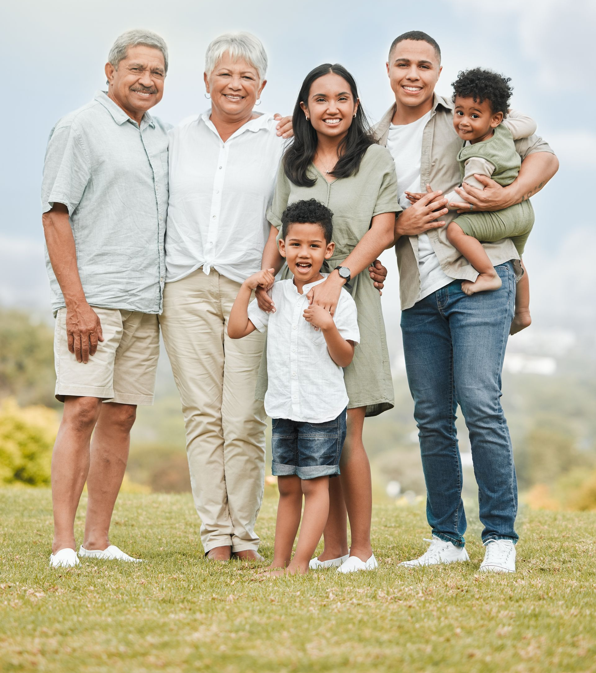 Family smiling outdoors, embracing. Two older adults, three adults, and two young children on a grassy hill.