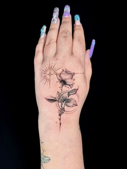 Hand with a black and white rose tattoo, a geometric sun, and long, colorful nails against a black background.