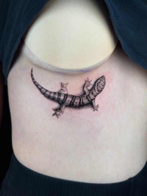 Tattoo of a detailed black and gray gecko on a person's side, next to a white bra strap.