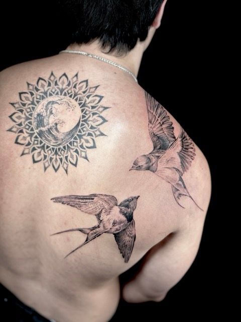Back tattoo of a sunburst design with a wave, and two swallows in flight; black background.