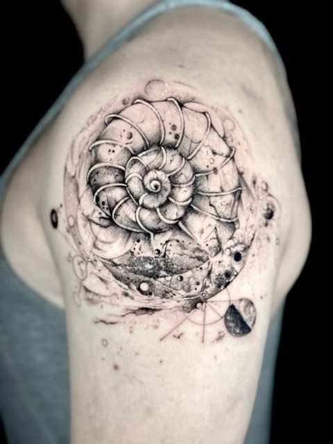 Tattoo of a nautilus shell on a shoulder, surrounded by nebulous designs and geometric shapes.