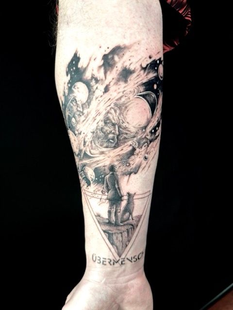 Tattoo of a figure and dog on a cliff, facing a cosmic face, above the word 