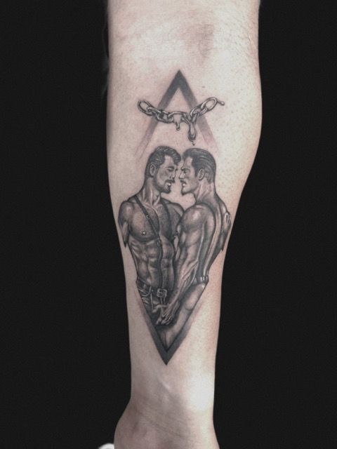 Tattoo on forearm: two muscular men, chain, triangle, leather suspenders, gray scale, black background.