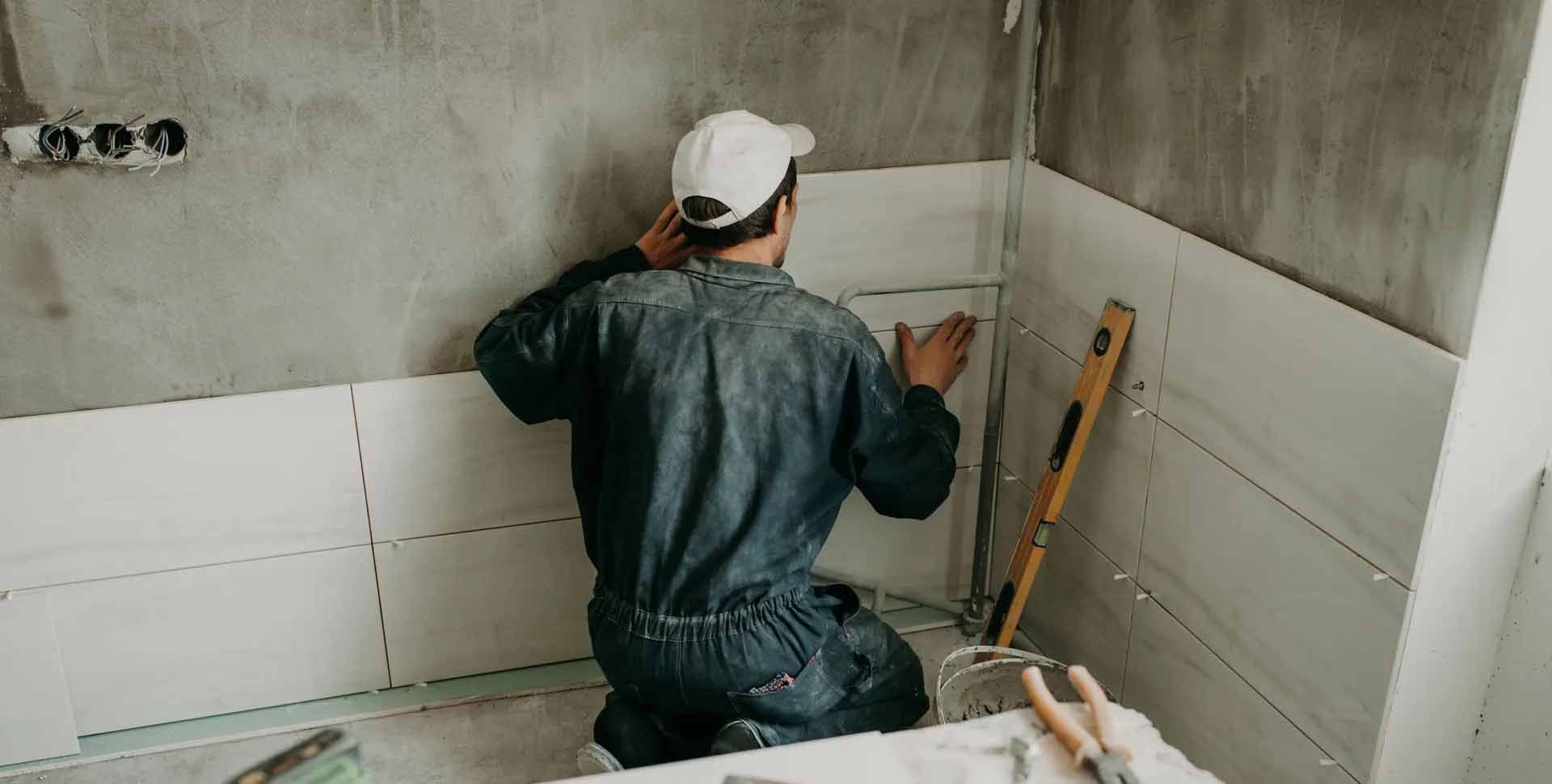 Homme en combinaison bleue et casquette blanche carrelant un mur dans une pièce en construction.