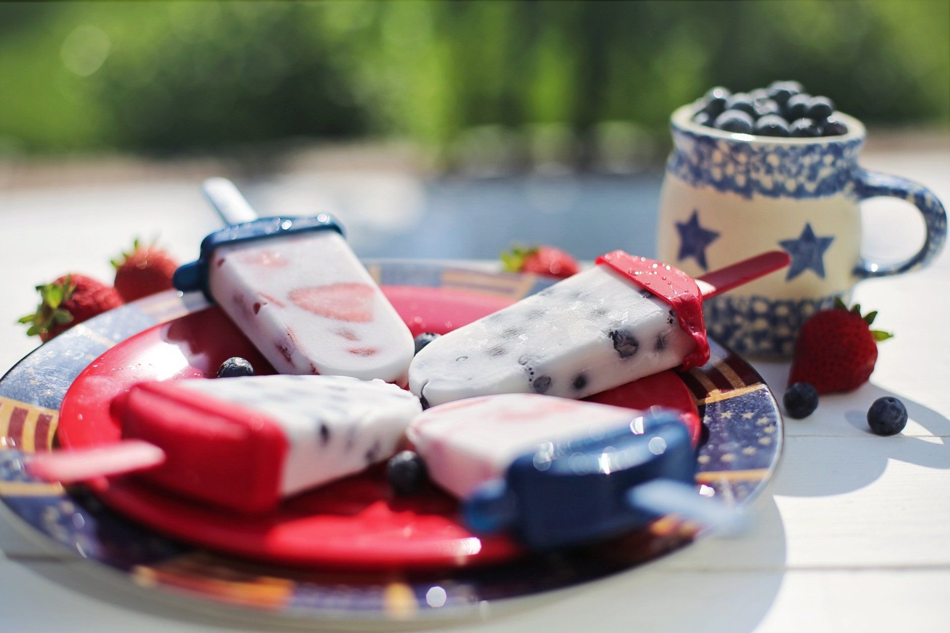 A plate of red white and blue popsicles with strawberries and blueberries on a table.