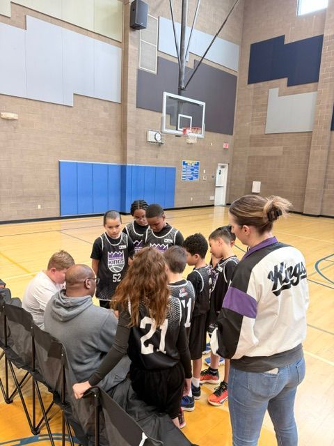 Basketball team huddled on a gym court during a timeout, with a coach speaking to them.