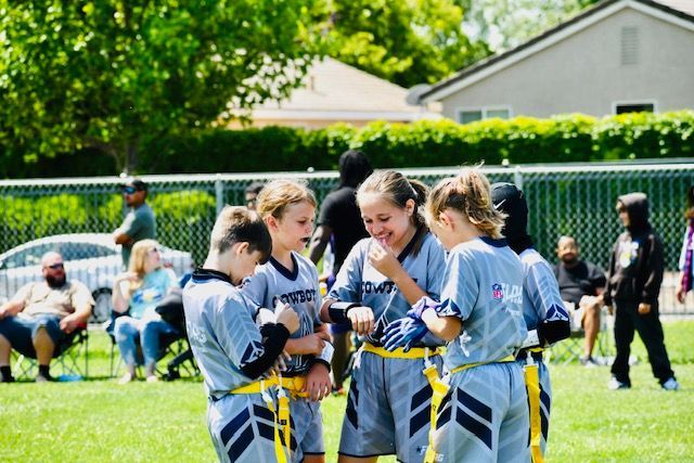 Girls in gray sports uniforms huddle on a sunny field, smiling and checking their gear before a game.