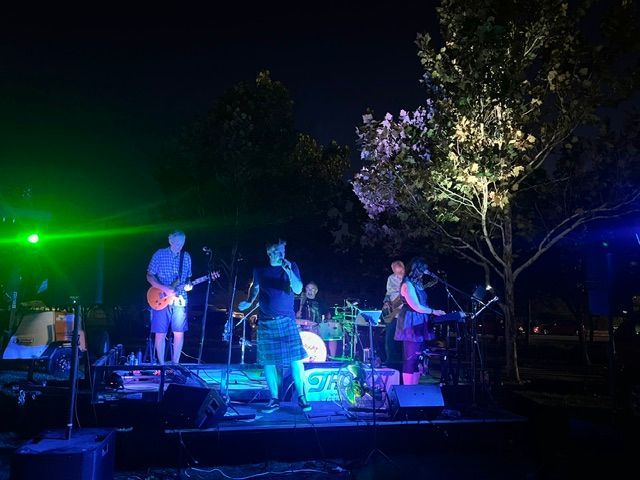 Band performing on a dark outdoor stage with blue and green lights, trees in the background.