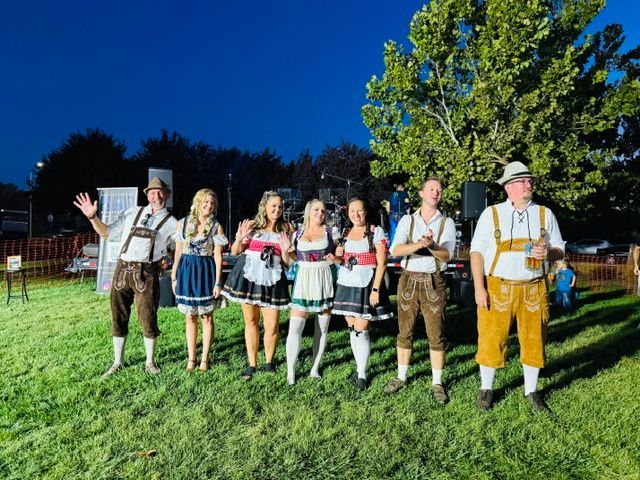 Group in traditional alpine clothing standing on grass at night, waving under a bright blue sky.