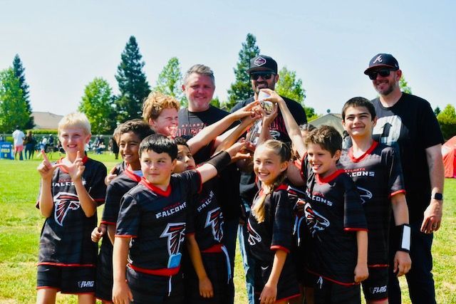 Youth football team and coaches posing together outdoors with a trophy under a blue sky