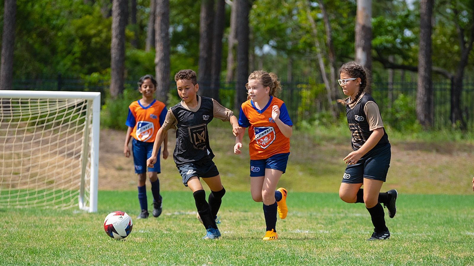 Youth soccer players in black and orange uniforms sprint toward the ball near the goal on a grassy field