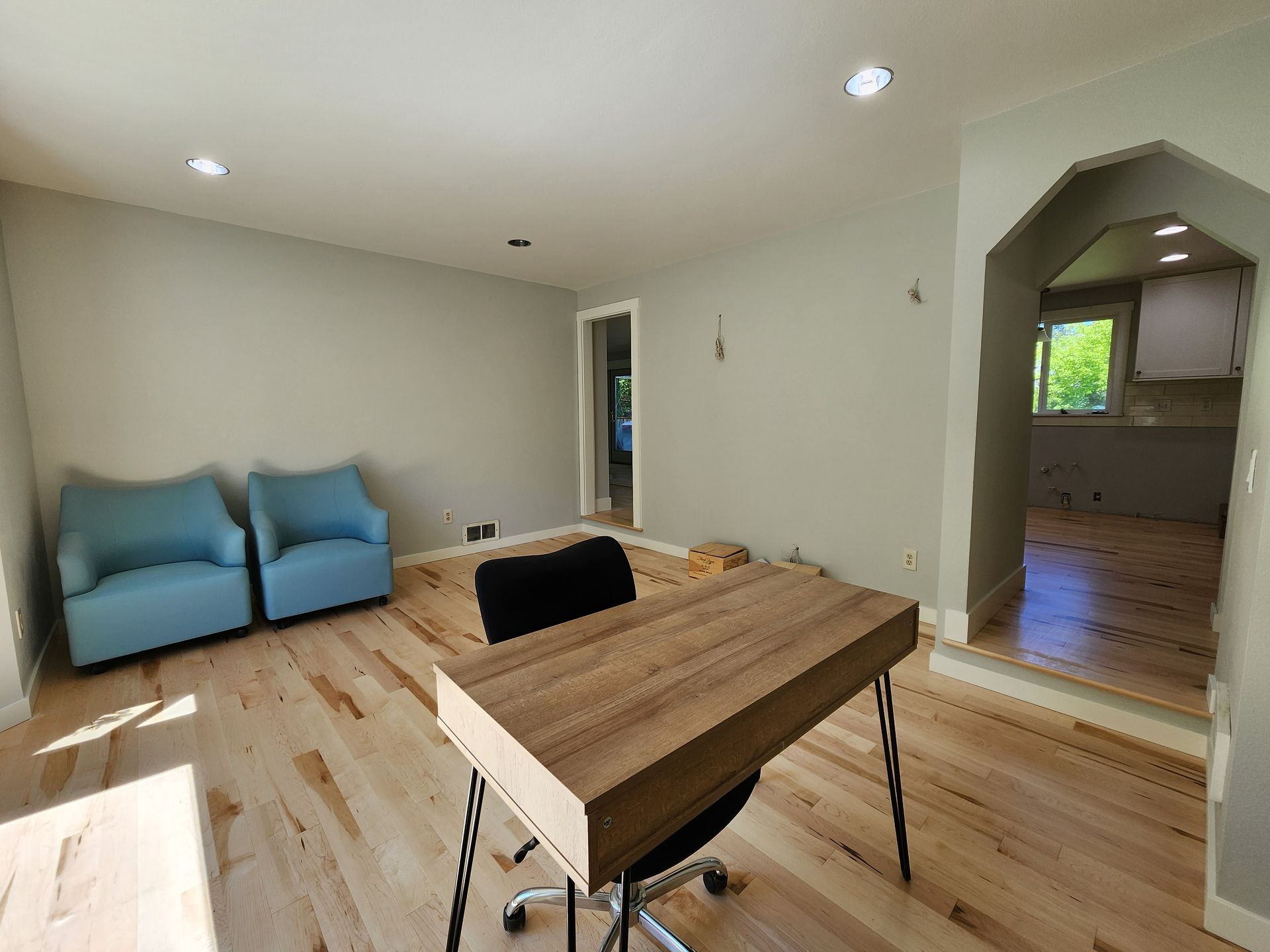 Room with light wood floor, gray walls, blue chairs, wooden desk, and a doorway to a kitchen.
