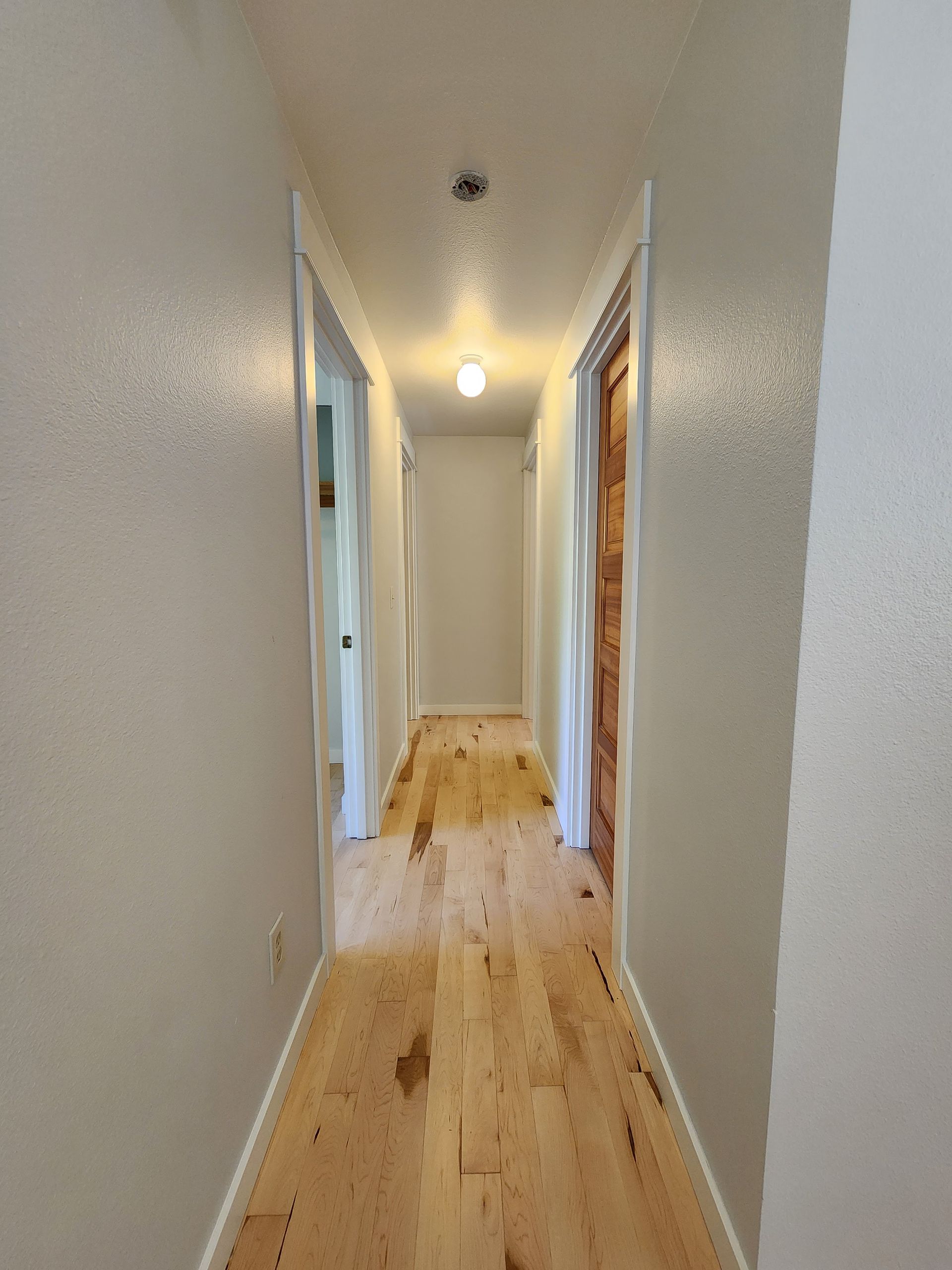Narrow hallway with wood floor, white walls and ceiling, doors on both sides, light at the end.