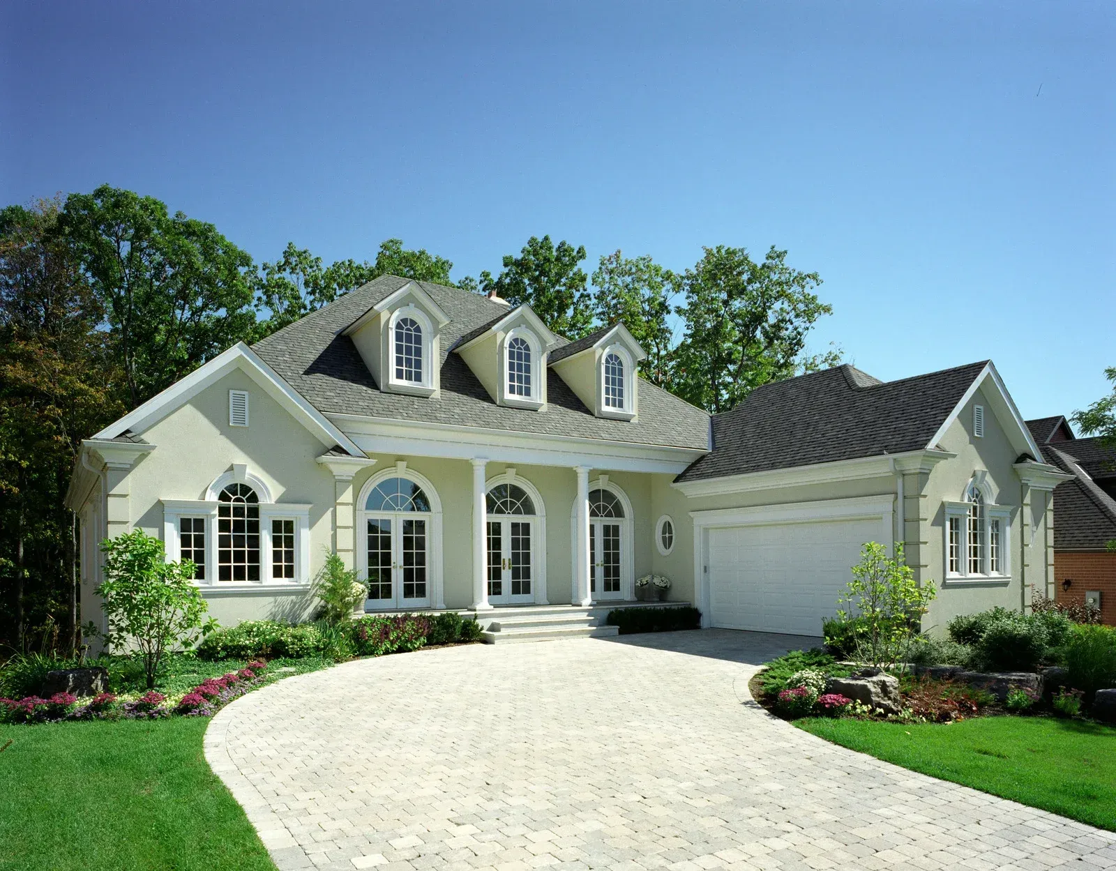 Elegant beige house with white trim, arched windows, and a winding stone driveway on a sunny day.