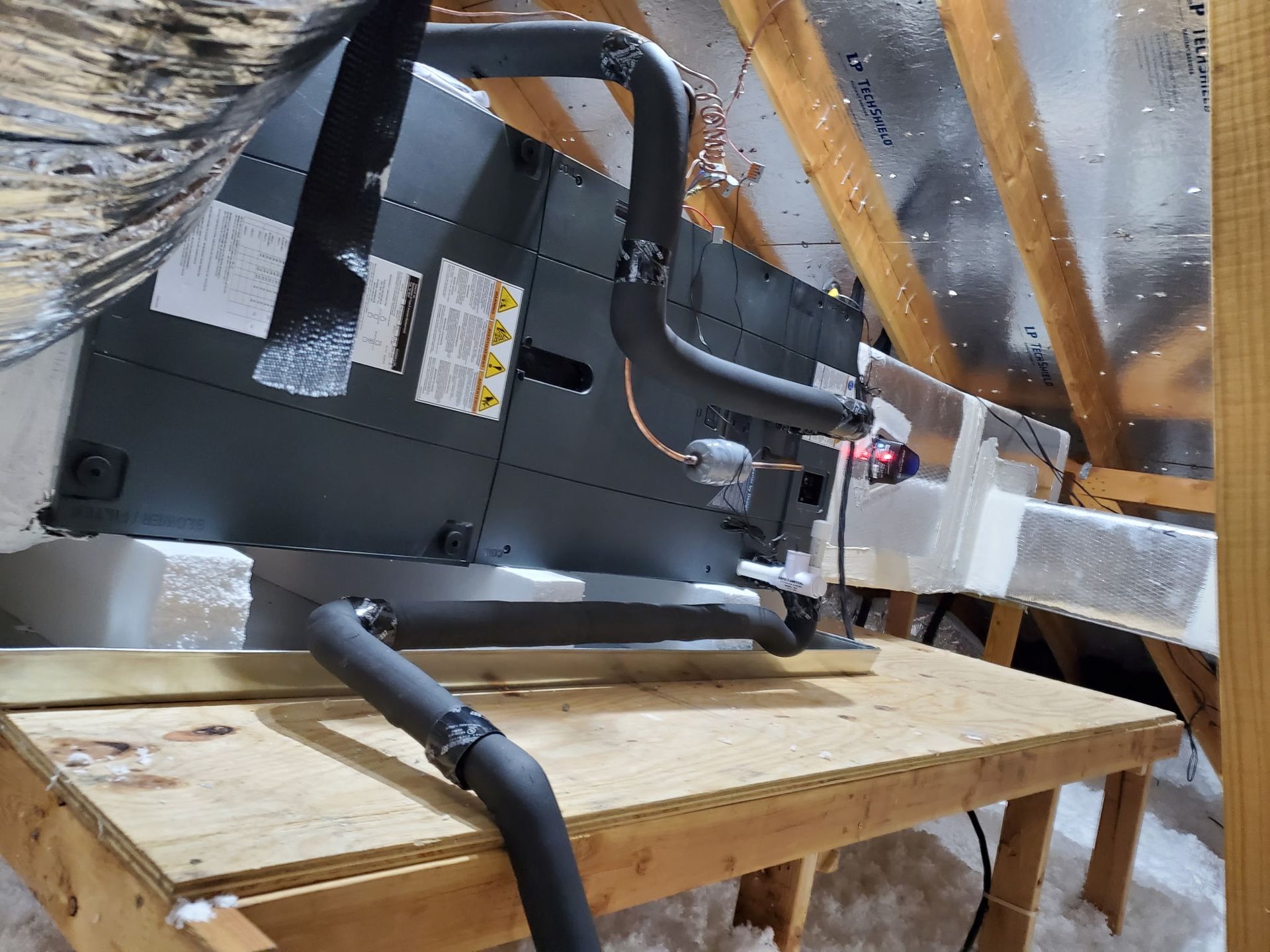 HVAC unit in an attic. Gray and black with insulated pipes on a wooden platform.