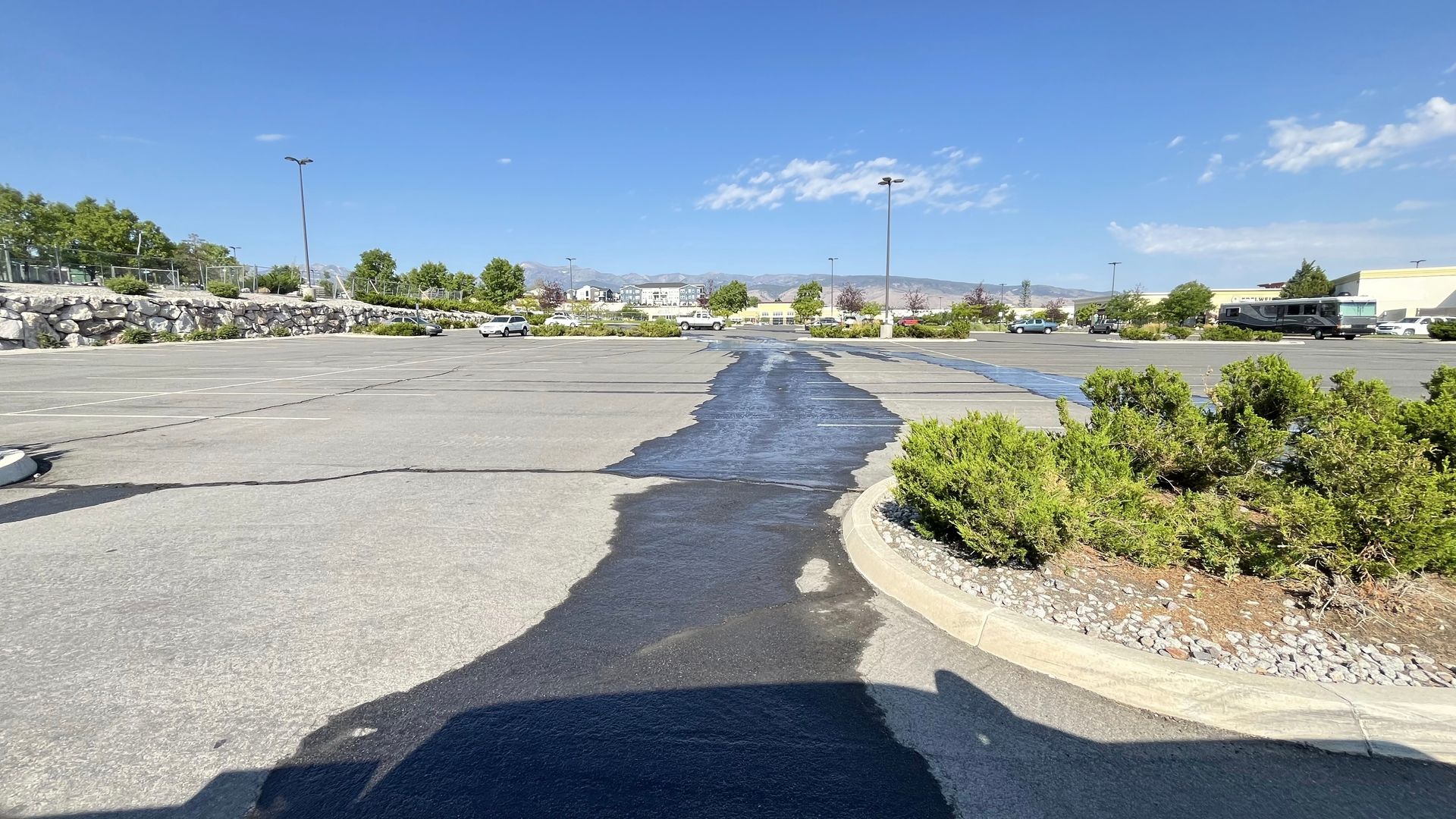 Asphalt parking lot with dark water pooling. Blue sky, sparse vegetation, distant buildings.