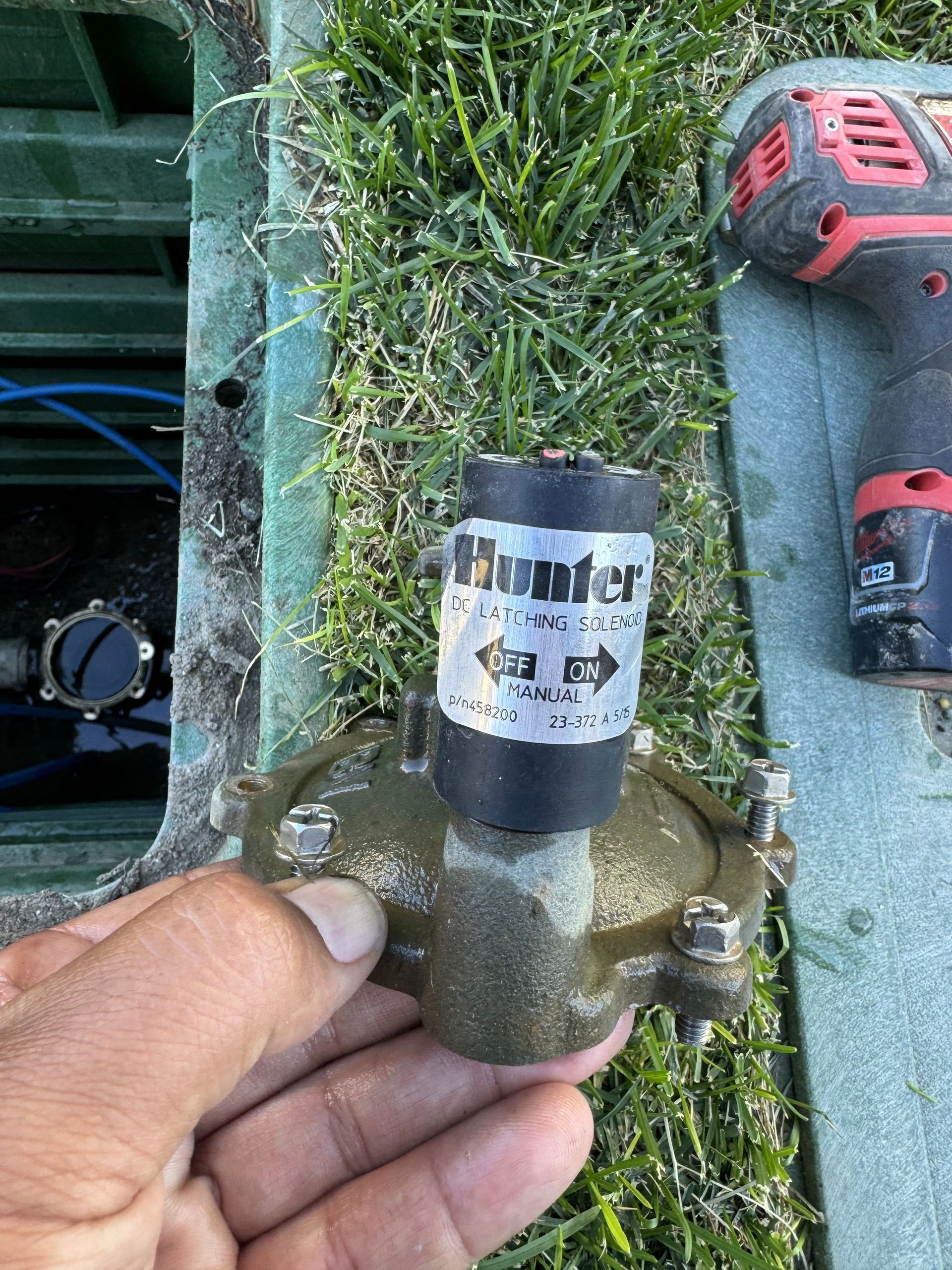 Hand holding a Hunter irrigation valve removed from a green plastic box on grass. A power drill is in the frame.