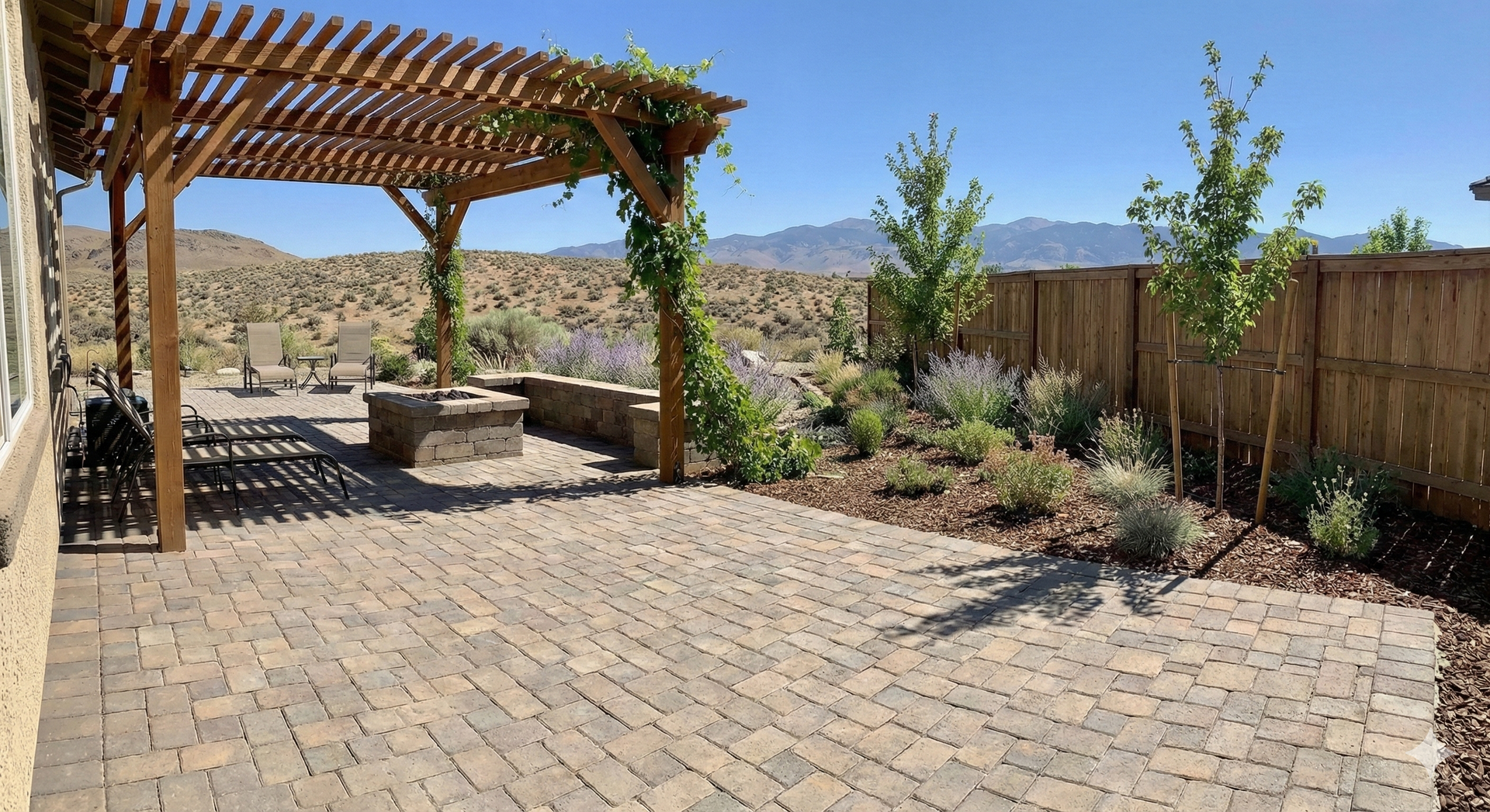 Patio with pergola, stone pavers, fire pit, and landscaping, against a desert backdrop under a blue sky.