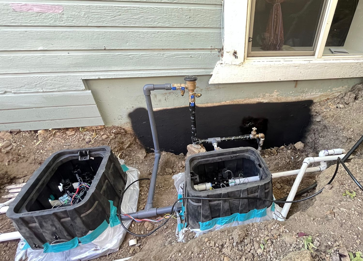 Irrigation system components, including control boxes, piping, and a water supply, are installed near a building's foundation.