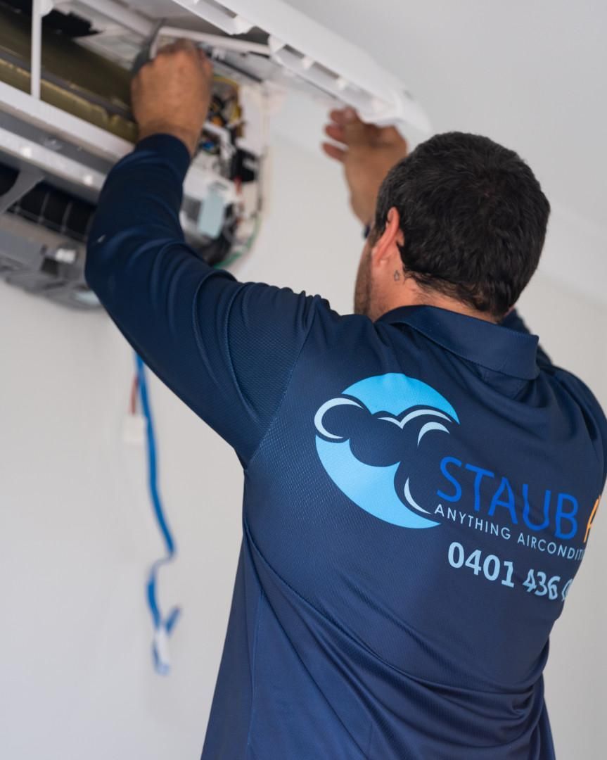 A Man Is Working on An Air Conditioner with A Staub Shirt On — Staub Air in Garbutt, QLD