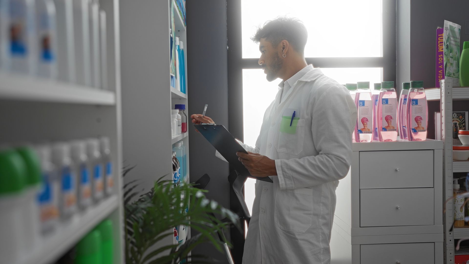 Una persona con bata blanca de laboratorio escribe en un portapapeles en el pasillo de una farmacia