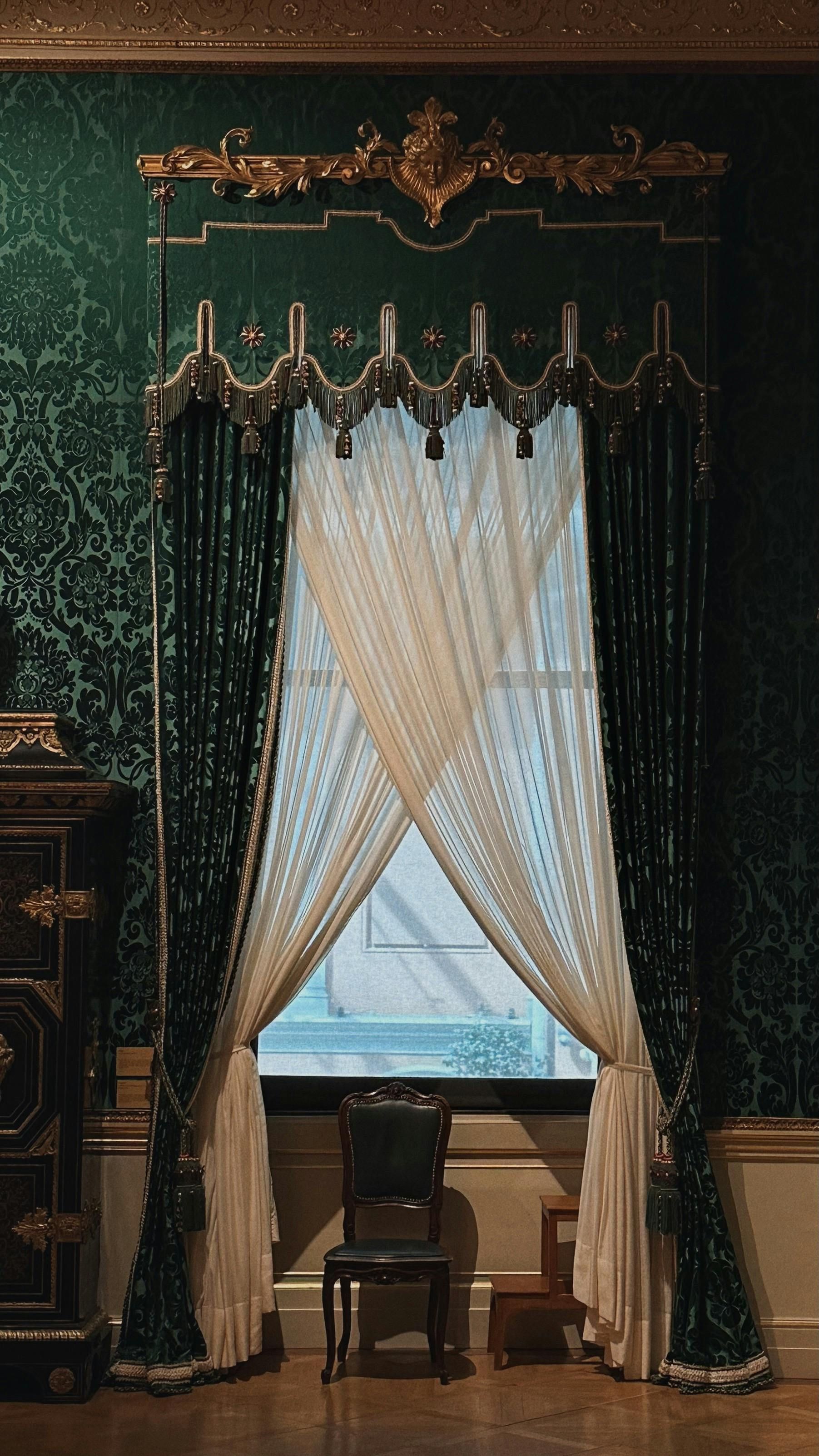 Ornate green and gold curtains frame a window, with a white lace sheer, a black chair, and a small wooden step stool.