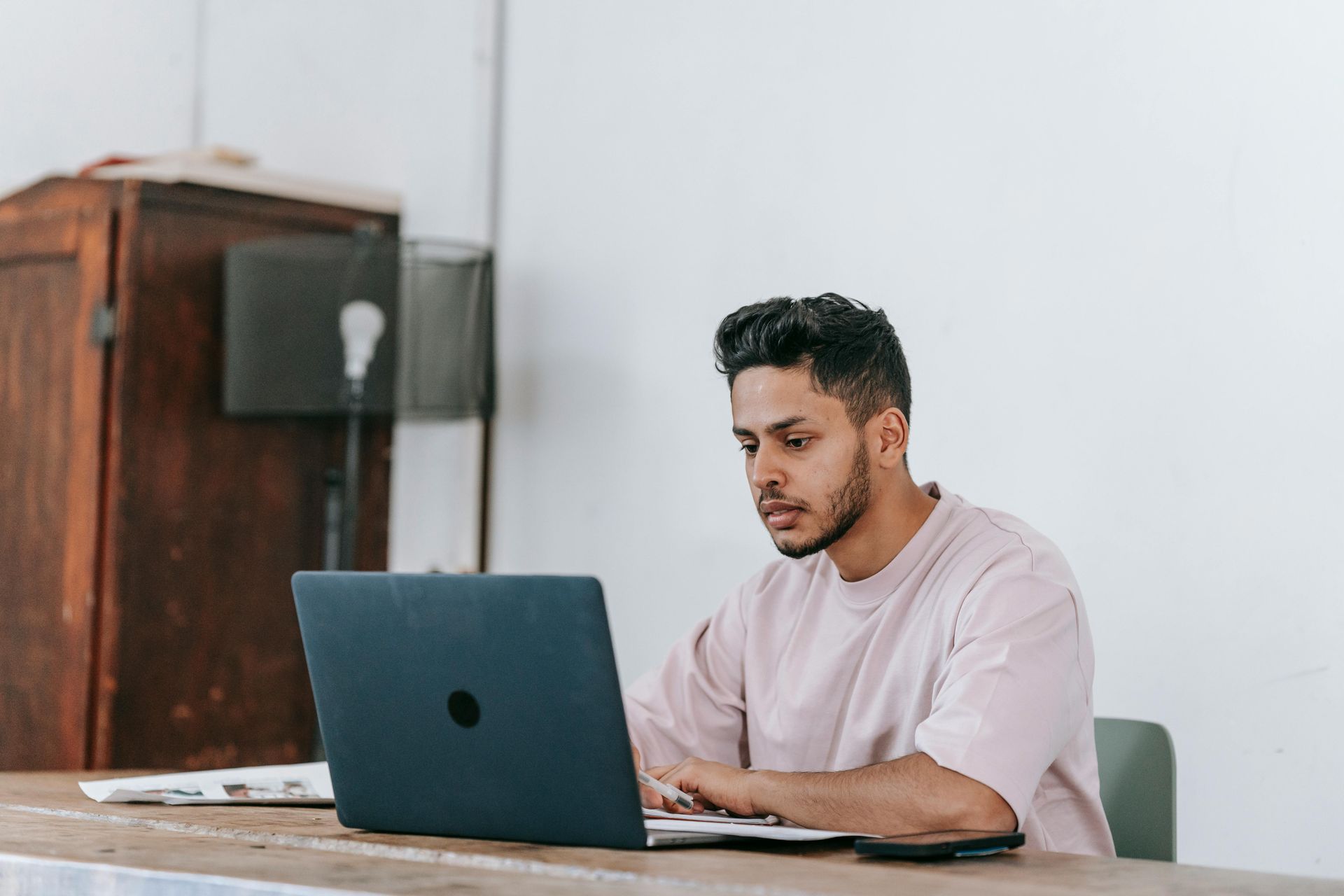Man in pink shirt typing on laptop at wooden desk. White wall and brown cabinet in background.