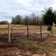 Rural metal gate in a fenced field with muddy ground, trees, and cloudy sky