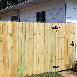 Wooden fence gate with black hinges in front of a white house exterior.