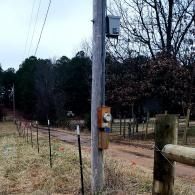 Wooden utility pole with call box beside a rural dirt road and fence, overcast wooded background