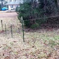 Woods edge with a wire fence and black posts beside a grassy yard near a house