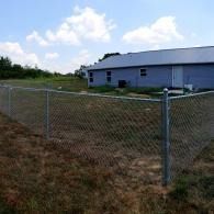 Chain-link fence around a grassy yard beside a blue industrial building under a partly cloudy sky