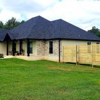 Single-story house with dark roof, light brick walls, and a wooden fence beside a green lawn.
