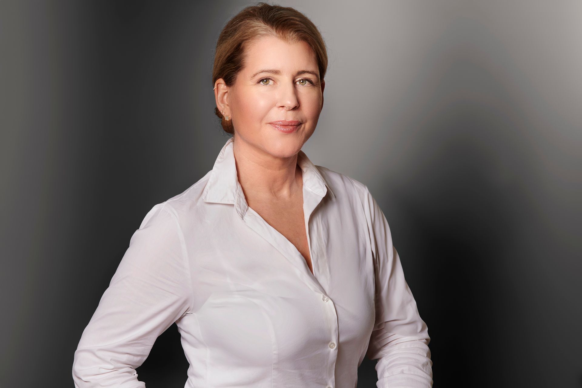 Eine Frau in einem weißen Hemd steht mit verschränkten Armen vor einem Fenster.